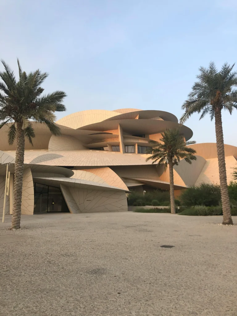 Image shows the exterior of the National Museum of Qatar on a pleasant day with two palm trees on either side of the building.