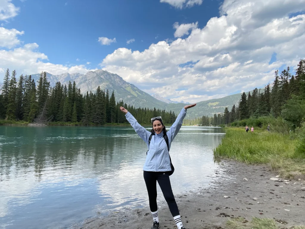 blogger standing with her arms raised in front of beautiful scenery of mountains, lake and trees against a cloudy blue sky in Banff, Alberta, Canada