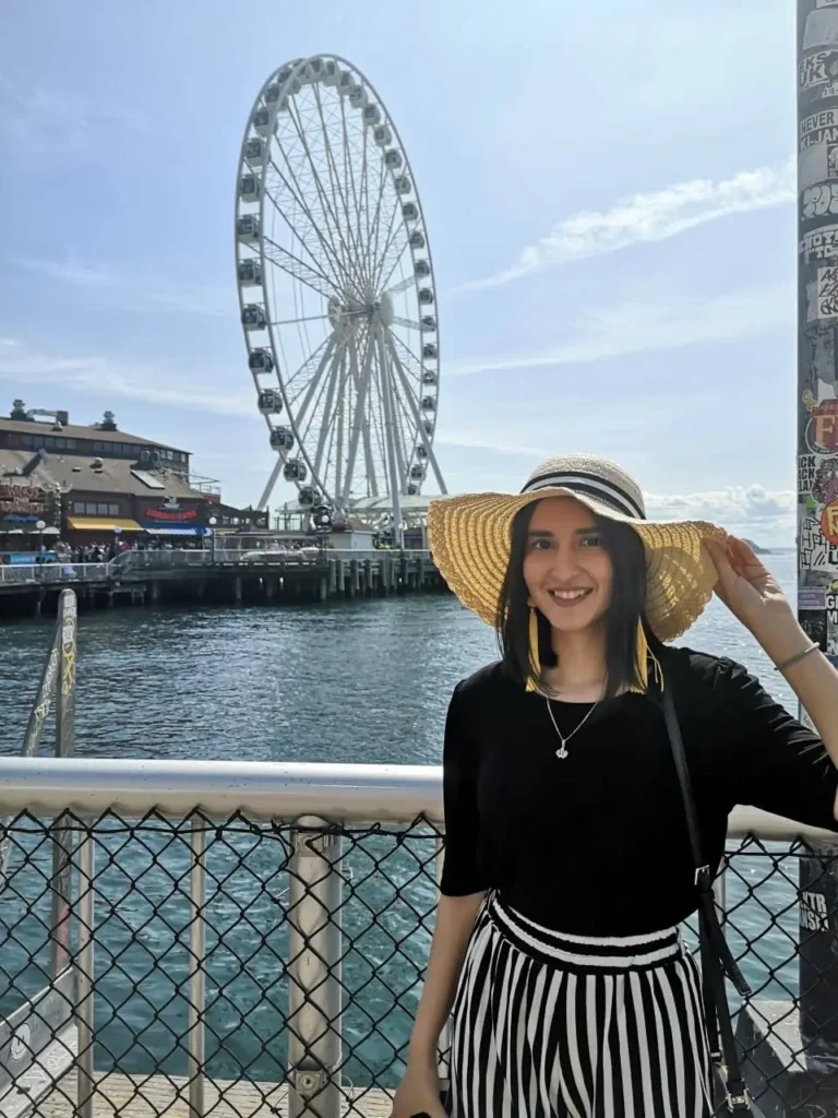 BLOGGER STANDING WITH A SUN HAT IN FRONT OF the Seattle pier with the Ferris wheel and water in the background