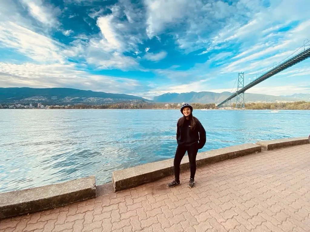 Blogger is standing and smiling in front of a beautiful scenery featuring cloudy blue skies, a bridge and mountains in Vancouver, BC, Canada